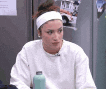 a woman wearing a headband and a white sweater is sitting in front of a refrigerator holding a water bottle .