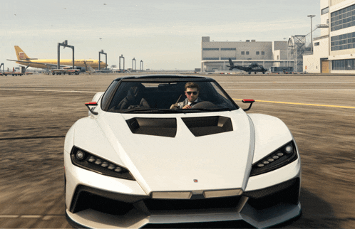 a man is driving a white sports car on a runway with a plane in the background