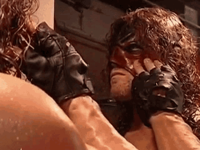 a wrestler wearing a mask and gloves is touching his face with his hands .