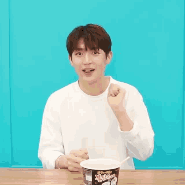 a young man is sitting at a table holding a cup of noodles and making a fist .