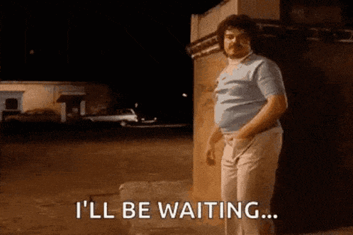 a man with a mustache is standing in front of a building and saying i 'll be waiting