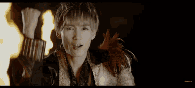 a man in a fur coat with feathers on his shoulder is standing in front of flames
