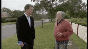 a man in a suit talks to another man in a red sweater with the number 9 on the sidewalk in front of them