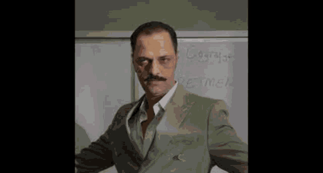 a man in a suit with a mustache is standing in front of a white board .