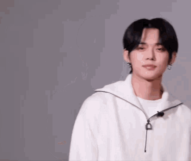 a young man with black hair and earrings is wearing a white sweater and looking at the camera .