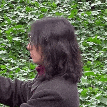 a woman wearing glasses stands in front of a lush green bush