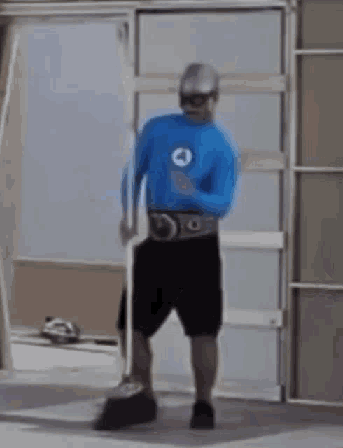 a man in a blue shirt and black shorts is standing in front of a door holding a stick .