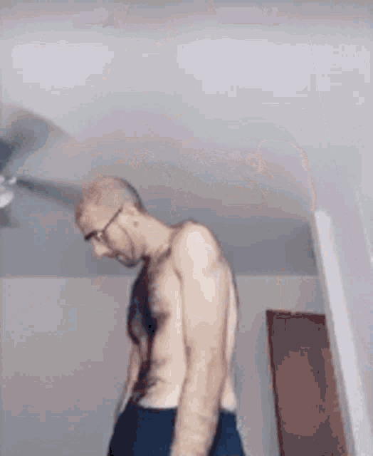 a shirtless man with glasses is standing in a room with a ceiling fan .