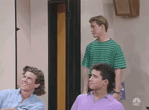 a group of young men are sitting in a room . one of the men is wearing a purple shirt .