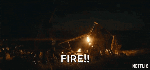 a group of people are standing in a field at night with a fire coming out of a catapult .