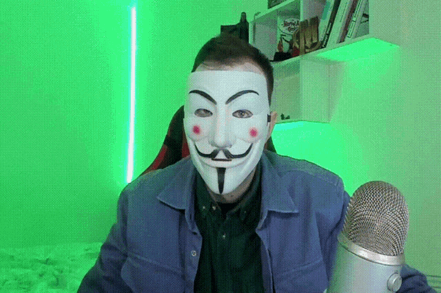 a man wearing a anonymous mask holds a microphone in front of a green background