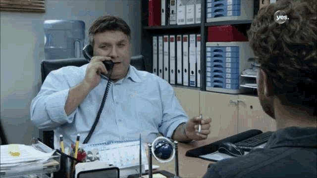 a man sitting at a desk talking on a phone with a yes logo on the bottom