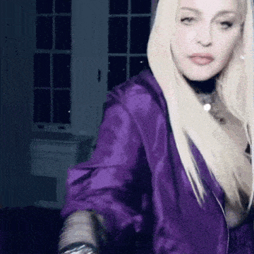 a woman with blonde hair is wearing a purple jacket and standing in front of a window .