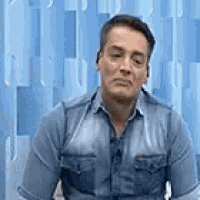 a man in a blue shirt is sitting in front of a blue wall and making a funny face .