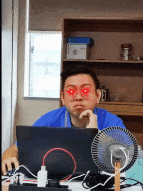 a man with red eyes is sitting at a desk with a laptop and a fan