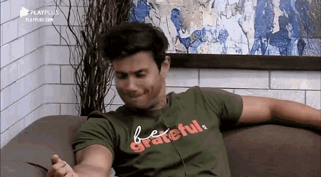 a man is sitting on a couch wearing a green t-shirt that says be grateful .