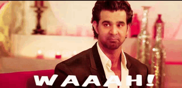 a man in a suit and white shirt is sitting on a red couch with the words waaah written on the bottom .
