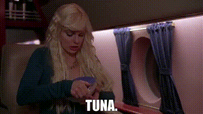 a woman in a wig is sitting on a plane with the word tuna on the screen .