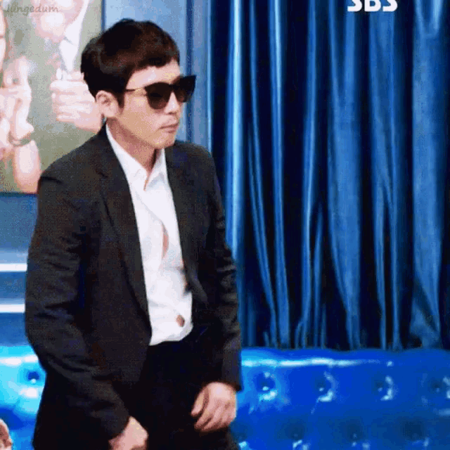 a man in a suit and sunglasses is standing in front of a blue curtain
