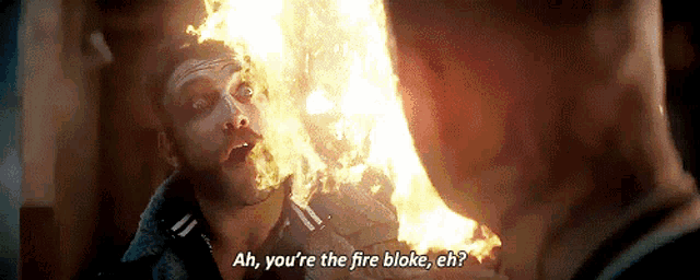 a man is standing in front of a fire and talking to another man .