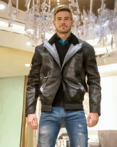 a man wearing a black leather jacket and jeans stands in front of a chandelier