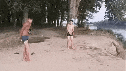 two men are standing on a sandy beach next to a river and trees .