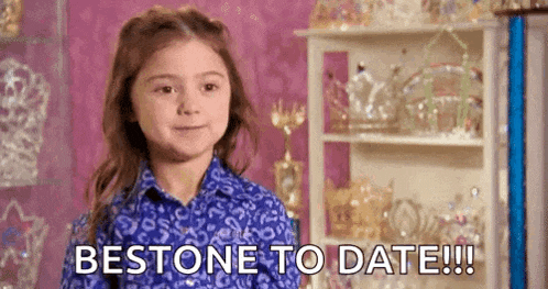 a little girl in a blue shirt is standing in front of a shelf and saying `` bestone to date '' .