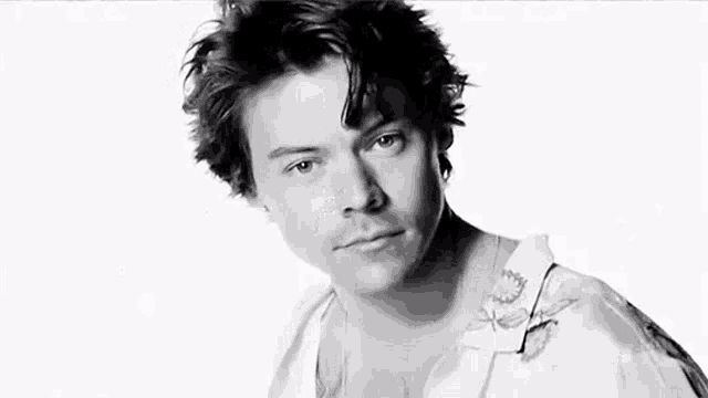 harry styles is wearing a white shirt with a tattoo on his chest and looking at the camera in a black and white photo .