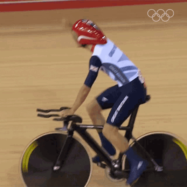 a british athlete is riding a bike on a track with the olympic rings in the background
