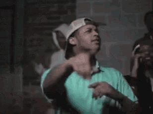 a man wearing a hat and a green shirt is standing in a dark room with his fist in the air .