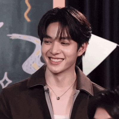 a young man wearing a necklace and a brown jacket is smiling .