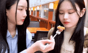 two girls are sitting at a table eating food from a fork