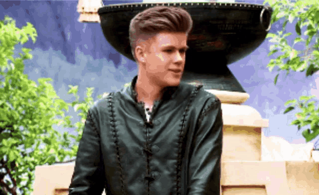 a young man in a green leather jacket stands in front of a fountain .