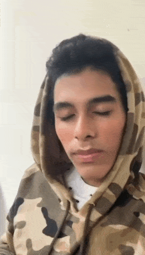 a young man wearing a camouflage hoodie with his eyes closed is looking down .