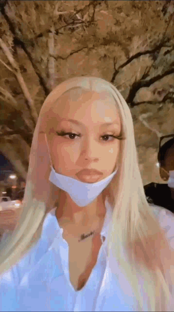 a woman wearing a face mask and a white shirt is standing in front of a tree .