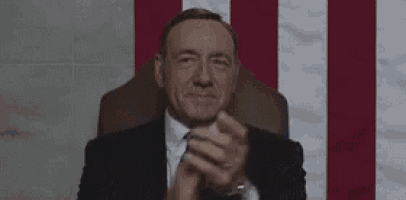 a man in a suit and tie is sitting in front of a flag and clapping his hands .