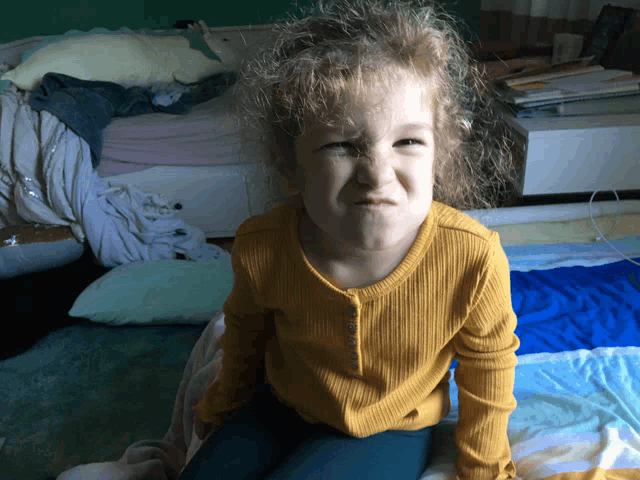 a little girl in a yellow shirt is sitting on a bed making a funny face