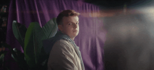 a young man with red hair is standing in front of a purple curtain .