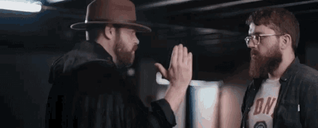 two men are giving each other a high five in a dark room . one of the men is wearing a hat and glasses .