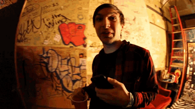 a man holding a cell phone in front of a wall with graffiti that says " to the wind "