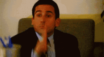 a man in a suit and tie is sitting on a couch and making a shhh gesture .