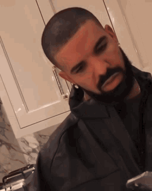 a man with a beard is wearing a black jacket and earrings while sitting in a kitchen .