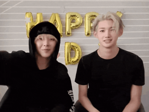 two young men sitting in front of a wall with balloons that say happy