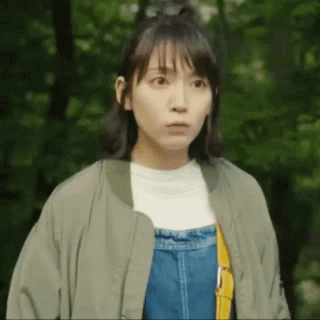 a woman in overalls and a bomber jacket is standing in the woods .