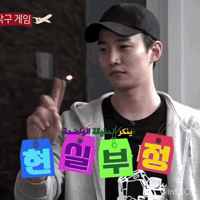 a man wearing a black hat holds up a peace sign while holding a colorful tag with korean writing