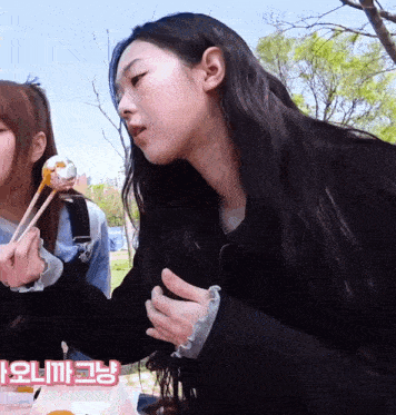 a girl with long black hair is holding a piece of food with chopsticks while another girl looks on