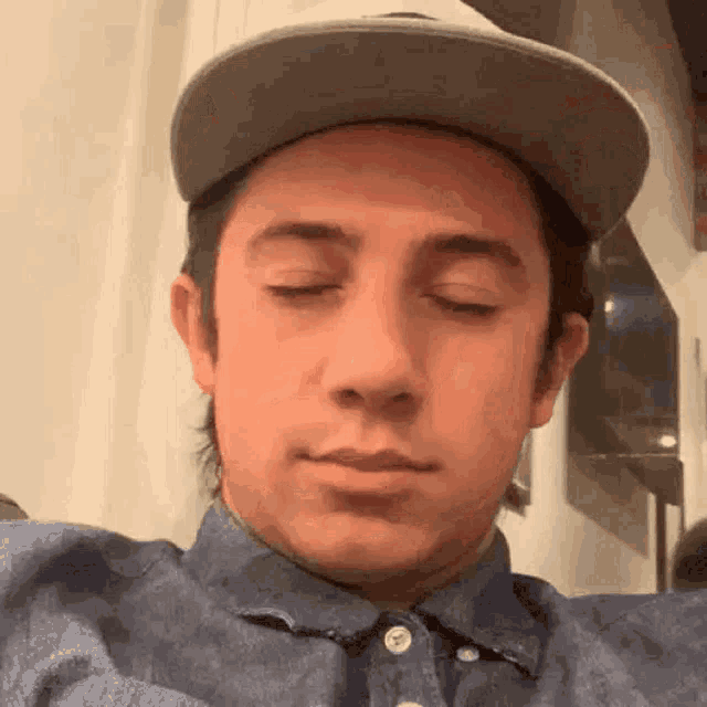 a man wearing a hat and a denim shirt is making a funny face with his eyes closed .