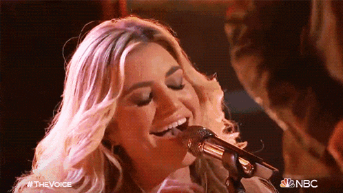 a woman is singing into a microphone with the nbc logo in the background .