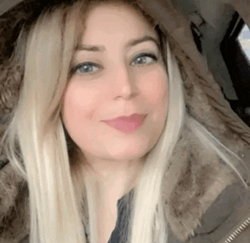 a woman with blonde hair and blue eyes is wearing a fur hooded jacket and smiling for the camera .