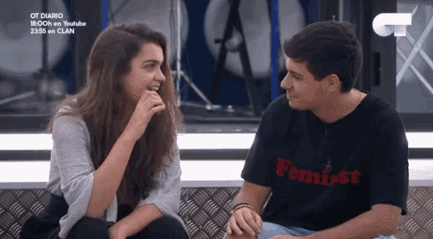 a man wearing a feminist shirt sits next to a woman laughing